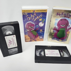 May include: Two Barney VHS tapes and their corresponding cases. One case is titled Barney in Outer Space, featuring a purple dinosaur in a space suit. The other case is titled Sing & Dance with Barney, featuring a purple dinosaur on a gold background.