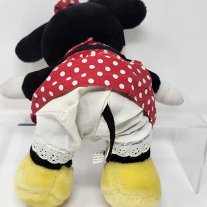 May include: A black and white plush toy of Minnie Mouse wearing a red dress with white polka dots and a white petticoat with lace trim. She has a red bow on her head and yellow shoes.