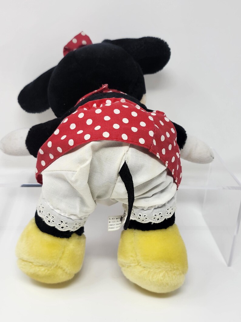 May include: A black and white plush toy of Minnie Mouse wearing a red dress with white polka dots and a white petticoat with lace trim. She has a red bow on her head and yellow shoes.