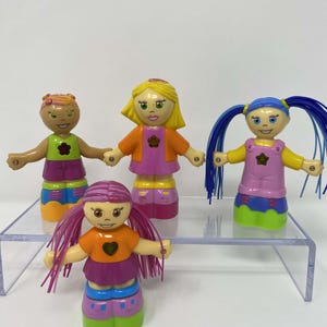 May include: Four colorful, stackable doll figures with various hair colors and outfits. Each doll has a smiling face and outstretched arms. The dolls are wearing tops, skirts, and shoes in shades of orange, pink, green, and blue. The dolls are standing on a clear, elevated surface.