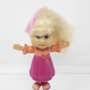 Peut inclure: Une petite poupée en plastique aux cheveux blonds avec une mèche rose. Elle porte une robe rose avec un corsage orange et un nœud. La poupée a les bras tendus sur les côtés.