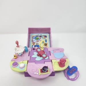 May include: A Little Pet Shop playset in shades of purple, pink, and green. The playset features a small pet figure, a miniature sink, and other accessories. The playset is open, revealing a detailed interior with various play areas.