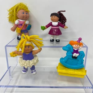 Peut inclure: Quatre figurines en plastique Cabbage Patch Kids. L'une a des cheveux jaunes, un haut rose avec le texte CPK et un short bleu. Une autre a des cheveux foncés, une robe violette et un col blanc. Une autre porte une tenue violette et un tutu. Une est sur un cheval à bascule bleu.