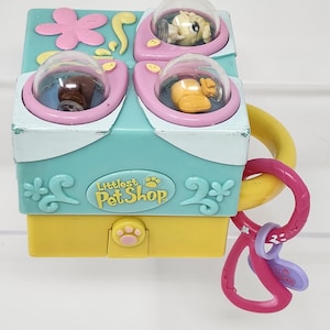 May include: A colorful Littlest Pet Shop toy featuring a teal, pink, and yellow design. The toy has four clear domes with small animal figures inside. A pink and purple clip is attached to the side. The toy has the words "Littlest Pet Shop" on it.