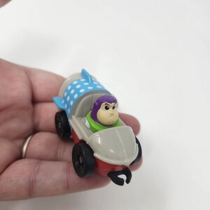 K&ouml;nnte beinhalten: Ein kleines Spielzeugauto mit Buzz Lightyear aus Toy Story. Das Auto hat einen roten und wei&szlig;en K&ouml;rper mit schwarzen R&auml;dern. Buzz Lightyear sitzt darin und tr&auml;gt seinen lila-gr&uuml;nen Raumanzug.