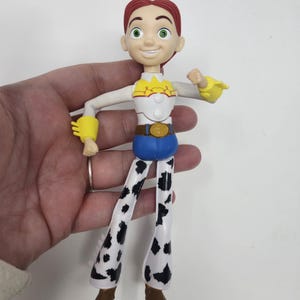 K&ouml;nnte beinhalten: Eine Jessie-Actionfigur aus dem Toy Story-Franchise. Die Figur hat rotes Haar, ein wei&szlig;es und gelbes Oberteil, blaue Hosen und Chaps mit Kuhmuster. Die Figur wird von einer Hand vor wei&szlig;em Hintergrund gehalten.