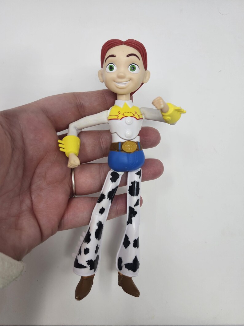 K&ouml;nnte beinhalten: Eine Jessie-Actionfigur aus dem Toy Story-Franchise. Die Figur hat rotes Haar, ein wei&szlig;es und gelbes Oberteil, blaue Hosen und Chaps mit Kuhmuster. Die Figur wird von einer Hand vor wei&szlig;em Hintergrund gehalten.