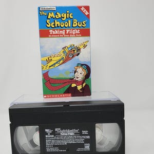 May include: A VHS tape of "The Magic School Bus: Taking Flight" with a colorful cover featuring a yellow school bus transformed into a plane. The tape is black and has the title printed on it.