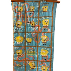 Op de afbeelding: Een lichtblauwe slaapzak met een herhalend rasterpatroon van bruine, bamboe-achtige lijnen en meerdere illustraties van SpongeBob SquarePants in verschillende houdingen. De tekst SpongeBob SquarePants is in het geel en groen op de stof gedrukt.