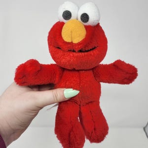 Può includere: Un peluche rosso di Elmo con grandi occhi bianchi, pupille nere e un naso giallo. Il personaggio di Sesame Street ha le braccia tese ed è fatto di materiale morbido e soffice. Un classico giocattolo per bambini.
