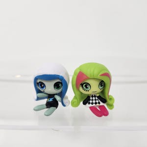 Peut inclure: Deux petites poupées stylisées avec de grands yeux. L'une a la peau bleue, une capuche blanche et une tenue noire avec un emblème bleu et jaune. L'autre a la peau verte, des cheveux verts avec des accents roses et une robe à carreaux noirs et blancs.