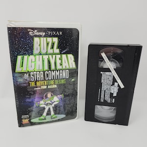 Peut inclure: Une cassette VHS Disney Pixar Buzz Lightyear of Star Command: The Adventure Begins avec sa pochette en carton d'origine. La pochette montre Buzz Lightyear sur un fond spatial, et la cassette en plastique noir porte une étiquette blanche.