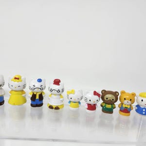 Puede incluir: Una colección de pequeñas y coloridas figuras que representan varios personajes. Las figuras incluyen un gato blanco con gafas, un vestido amarillo y un lazo rojo. Otros personajes incluyen un oso y un gato con un lazo rojo.