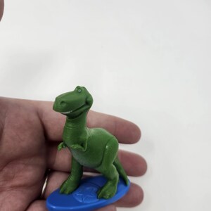 K&ouml;nnte beinhalten: Ein gr&uuml;nes Dinosaurier-Spielzeug aus Kunststoff auf einem blauen Surfbrett. Der Dinosaurier hat einen freundlichen Ausdruck und posiert wie beim Surfen. Das Surfbrett hat ein Stern-Design. Das Spielzeug ist ein Sammlerst&uuml;ck.