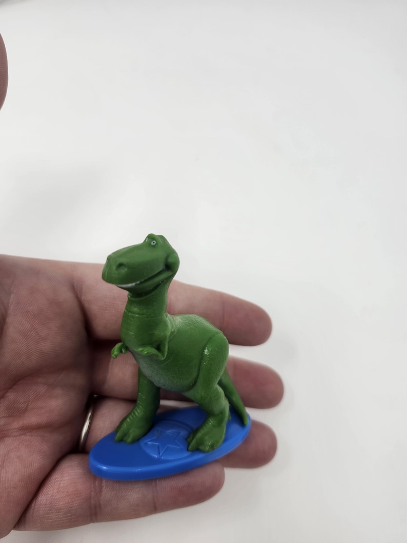 K&ouml;nnte beinhalten: Ein gr&uuml;nes Dinosaurier-Spielzeug aus Kunststoff auf einem blauen Surfbrett. Der Dinosaurier hat einen freundlichen Ausdruck und posiert wie beim Surfen. Das Surfbrett hat ein Stern-Design. Das Spielzeug ist ein Sammlerst&uuml;ck.