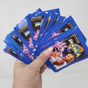 May include: A hand holding a fan of blue Space Jam sticker packs. Each pack features the Space Jam logo and cartoon characters. The packs are sealed and have the word "STICKERS" printed on them.