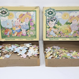 Peut inclure: Deux puzzles Cabbage Patch Kids vintage, chacun de 60 pièces, dans leurs boîtes d'origine. Les boîtes présentent des illustrations de Cabbage Patch Kids dans une scène extérieure ensoleillée. Les puzzles sont partiellement assemblés dans leurs plateaux en carton.