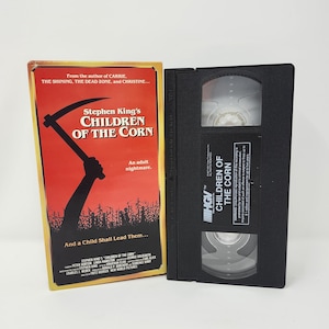 May include: A VHS tape and its case for the movie "Children of the Corn." The case is red and yellow with the movie title and a graphic of a scythe. The VHS tape is black with the movie title.