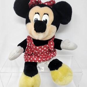May include: A plush Minnie Mouse toy wearing a red dress with white polka dots and a red bow with white polka dots. The toy has black ears, a black nose, and yellow shoes.
