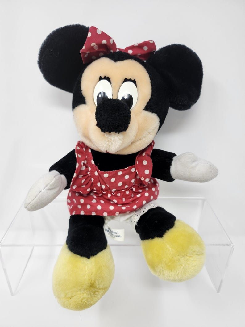May include: A plush Minnie Mouse toy wearing a red dress with white polka dots and a red bow with white polka dots. The toy has black ears, a black nose, and yellow shoes.