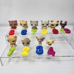 Puede incluir: Una colección de figuras de gatos en miniatura, coloridas y con diversos diseños. Cada figura está emparejada con un número del uno al siete en un color correspondiente. Los juguetes están dispuestos en una pantalla transparente de varios niveles.