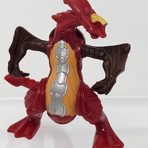Peut inclure: Figurine de dragon jouet rouge et marron avec une crête jaune sur la tête et un ventre segmenté orange et argenté. Le dragon a les bras et les jambes tendus, comme s'il était prêt à attaquer.