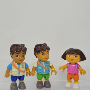 May include: Three plastic action figures of cartoon characters. Two figures are boys wearing vests and shorts. The third figure is a girl wearing a pink shirt and orange shorts. All figures have brown hair and are smiling.