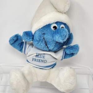 Puede incluir: Un muñeco de peluche de los Pitufos, con cuerpo azul, gorro blanco y pantalón blanco. El muñeco lleva una camisa blanca y azul con el texto "TRUE BLUE FRIEND". El Pitufo tiene una expresión sonriente y está sentado.