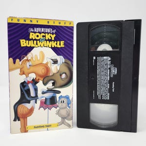 May include: A vintage VHS tape and case for "The Adventures of Rocky and Bullwinkle: Painting Theft." The case has a purple and yellow design with cartoon characters. A black VHS tape is next to the case.