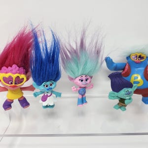 Puede incluir: Una colección de figuras de Trolls coloridos con cabello vibrante en tonos rosa, azul y verde. Las figuras tienen varios atuendos y accesorios, incluyendo gafas y una letra "B" en el pecho. Los juguetes se exhiben sobre una superficie transparente.