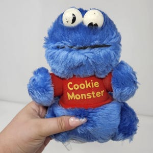 May include: A blue plush Cookie Monster toy with large white eyes and a red shirt. The shirt has the words "Cookie Monster" printed in yellow. The toy is soft and fluffy, with a smiling mouth.