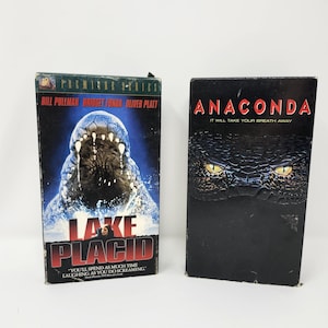 Peut inclure: Deux cassettes VHS. L'une présente une image bleue et blanche d'un visage de créature avec le texte "LAKE PLACID" et la citation "YOU'LL SPEND AS MUCH TIME LAUGHING AS YOU DO SCREAMING." L'autre cassette est noire avec le titre "ANACONDA" en rouge.