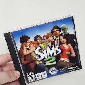 Peut inclure: Jeu vidéo The Sims 2 pour Mac dans un boîtier DVD en plastique transparent. La jaquette présente un groupe de personnages variés, dont un extraterrestre vert, avec le titre du jeu en bleu et vert. Le boîtier porte le logo EA et l'évaluation Teen.