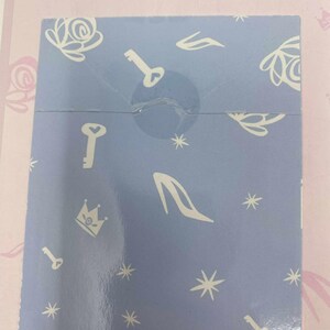 May include: A light blue box with a repeating pattern of white keys, high heels, crowns, and star shapes. The box is sealed with clear tape and sits on a pink surface with floral and crown designs.