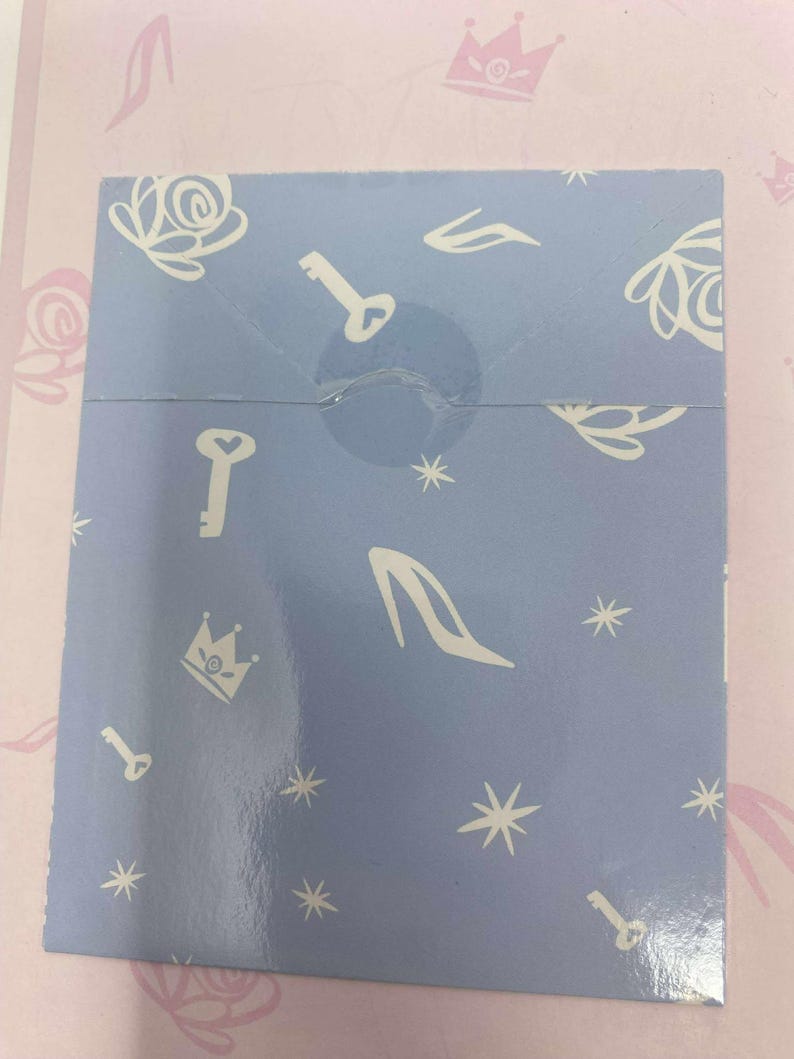 May include: A light blue box with a repeating pattern of white keys, high heels, crowns, and star shapes. The box is sealed with clear tape and sits on a pink surface with floral and crown designs.