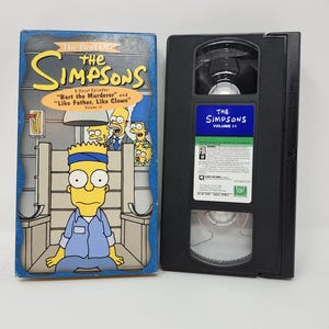 Può includere: Una videocassetta VHS e la sua custodia per "I Simpson" Volume 11. La custodia è blu con un'immagine a cartoni animati di Bart Simpson. Il nastro è nero con un'etichetta blu che dice "I Simpson Volume 11". La custodia ha anche il testo "The Best Of The Simpsons".