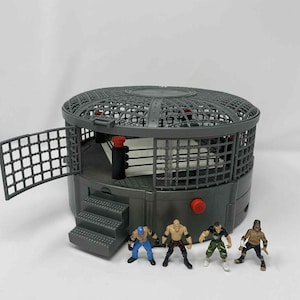 Peut inclure: Un ensemble de jeu de cage de lutte grise avec une porte ouverte et des marches, comprenant quatre figurines de lutteurs miniatures. La cage a un design circulaire avec un dessus grillagé et un bouton rouge. Les figurines sont dans diverses poses de lutte.