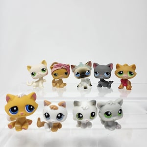 May include: A collection of nine small, colorful cat figurines. The figurines are in various poses and colors, including white, gray, tan, and orange, with large, expressive eyes. Some cats have accessories like bows or collars.