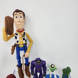 K&ouml;nnte beinhalten: Eine Sammlung von Toy Story-Charakter-Actionfiguren. Woody, in Cowboy-Kleidung, h&auml;lt ein Seil. Weitere Figuren sind ein lila Roboter, Buzz Lightyear, Jessie und ein gr&uuml;ner Dinosaurier. Die Spielzeuge sind aus Kunststoff und farbenfroh.