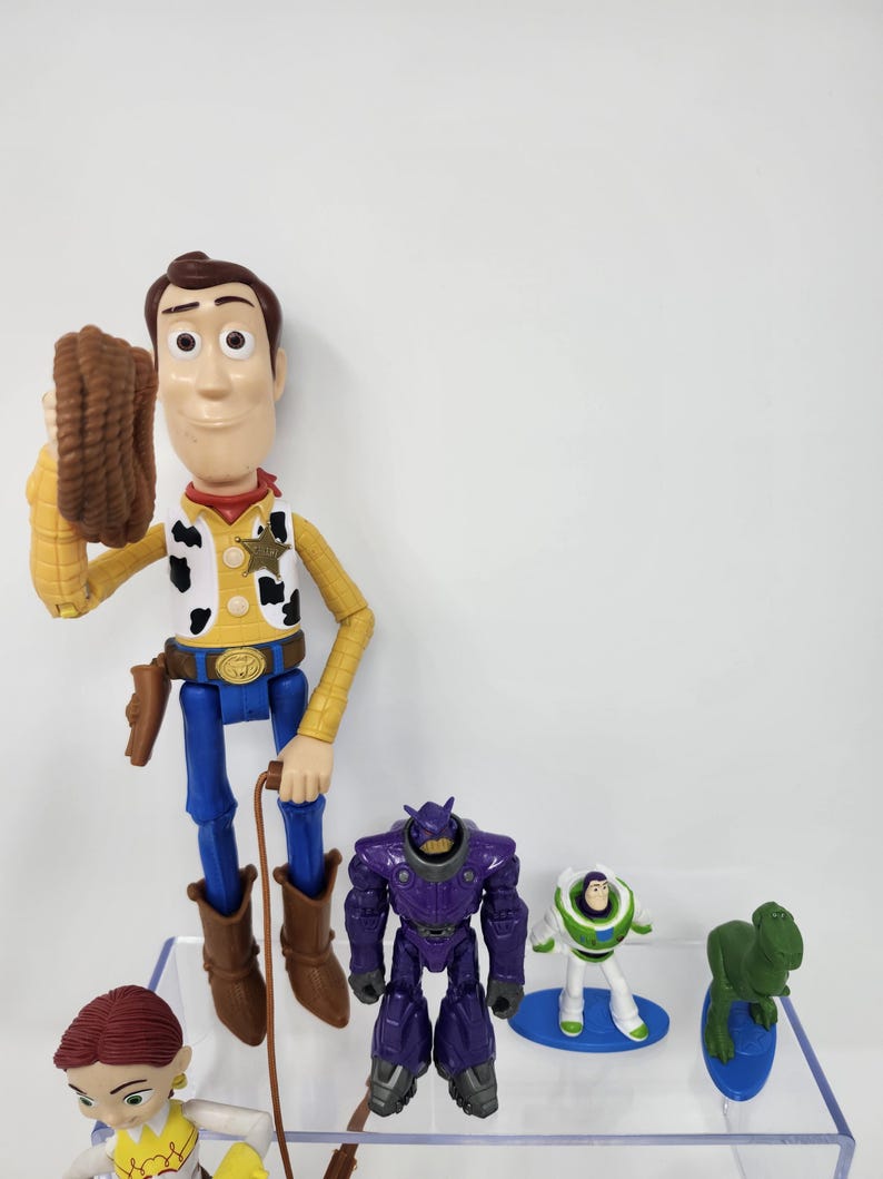 K&ouml;nnte beinhalten: Eine Sammlung von Toy Story-Charakter-Actionfiguren. Woody, in Cowboy-Kleidung, h&auml;lt ein Seil. Weitere Figuren sind ein lila Roboter, Buzz Lightyear, Jessie und ein gr&uuml;ner Dinosaurier. Die Spielzeuge sind aus Kunststoff und farbenfroh.