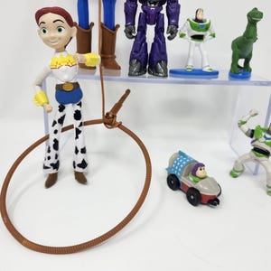 K&ouml;nnte beinhalten: Eine Sammlung von Toy Story-Charakterfiguren, darunter Jessie, Buzz Lightyear und Zurg. Jessie tr&auml;gt ein Cowboy-Outfit mit einem braunen Lasso. Buzz Lightyear tr&auml;gt einen gr&uuml;n-wei&szlig;en Raumanzug. Weitere Charaktere sind ein gr&uuml;ner Dinosaurier und ein Auto.