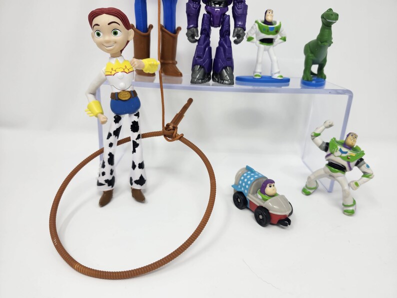 K&ouml;nnte beinhalten: Eine Sammlung von Toy Story-Charakterfiguren, darunter Jessie, Buzz Lightyear und Zurg. Jessie tr&auml;gt ein Cowboy-Outfit mit einem braunen Lasso. Buzz Lightyear tr&auml;gt einen gr&uuml;n-wei&szlig;en Raumanzug. Weitere Charaktere sind ein gr&uuml;ner Dinosaurier und ein Auto.