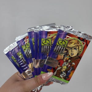 May include: A hand holding several sealed packs of "Jonny Quest" trading cards. The packs have a purple and silver foil design with illustrations of characters from the animated series. The text "JONNY QUEST" is visible on the packs.
