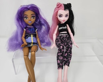 May include: Two fashion dolls with long purple and pink hair. The doll on the left has brown skin and is wearing a blue and yellow top, a denim skirt, and purple shoes. The doll on the right has pale skin and is wearing a black tank top with the word "FANTASY" on it, black and pink pants, and pink shoes.