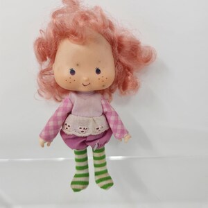 Peut inclure: Une poupée vintage avec un visage rond, des taches de rousseur et des cheveux bouclés roses. La poupée porte une chemise à manches longues rose et blanche à carreaux, une jupe rose et des chaussettes rayées vertes et blanches. La poupée a une expression souriante.