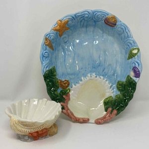 Puede incluir: Un juego de servir de cerámica con una fuente grande, con bordes festoneados y un cuenco más pequeño con forma de concha. La fuente grande tiene un interior azul con detalles de olas y está adornada con estrellas de mar y conchas. El cuenco más pequeño es blanco con un exterior texturizado en forma de concha.