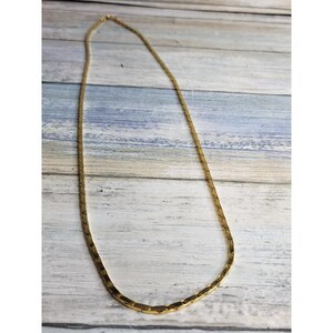 May include: A gold-toned necklace with a flat, geometric chain design. The necklace is displayed against a light blue and white wooden background. The chain has a subtle, textured appearance, and the clasp is visible at the top.