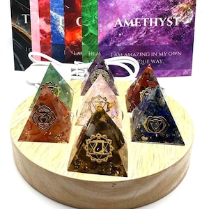 May include: Seven chakra crystal pyramids on a wooden base with a white cord. Each pyramid is a different color and has a gold symbol on it. The card in the background says "Amethyst, I am amazing in my own way."