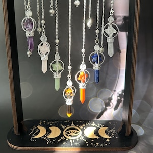 May include: A wooden stand with a black base and gold accents displays eight crystal pendulums. The pendulums are suspended from silver chains and feature various colored crystals, including amethyst, clear quartz, green aventurine, orange carnelian, red jasper, and blue lapis lazuli.