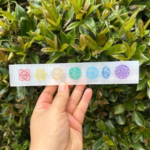 May include: A clear crystal charging plate with seven chakra symbols etched in rainbow colors. The symbols are arranged in a row from red to purple.